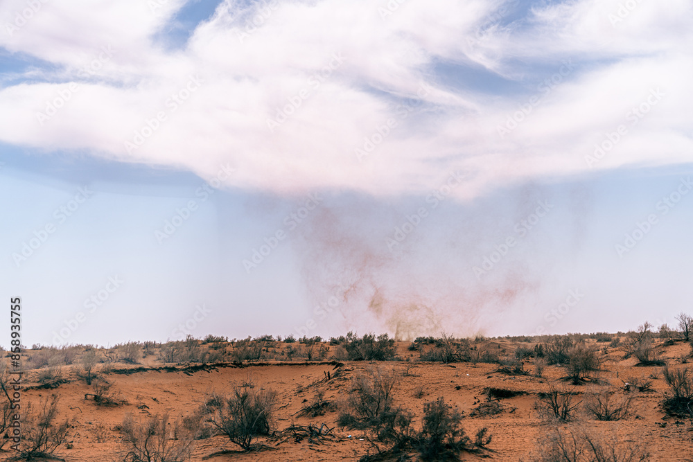 Fototapeta premium A cloud of dust rises from the desert floor. The sky is clear and blue, with a few clouds scattered throughout. The scene is desolate and barren, with only a few sparse trees in the distance