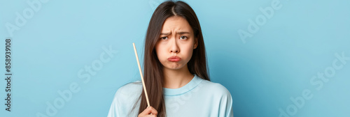 Young woman with a pouty expression holding a chopstick, standing against a light blue background, capturing a playful and slightly annoyed mood.