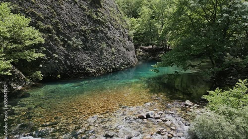 An aerial view of the Voidomatis river winding through the Vikos Gorge in Greece. The water is a vibrant turquoise color, contrasting with the green foliage and rocky cliffs.