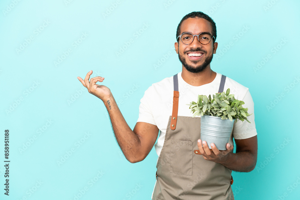 Gardener latin man holding a plant isolated on blue background extending hands to the side for inviting to come Gardener latin man holding a plant isolated on blue background extending hands to the side for inviting to come