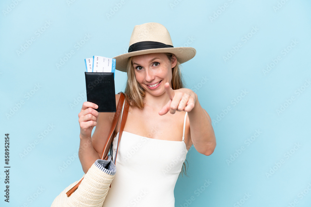Young blonde woman in swimsuit holding passport isolated on blue background points finger at you with a confident expression Young blonde woman in swimsuit holding passport isolated on blue background points finger at you with a confident expression