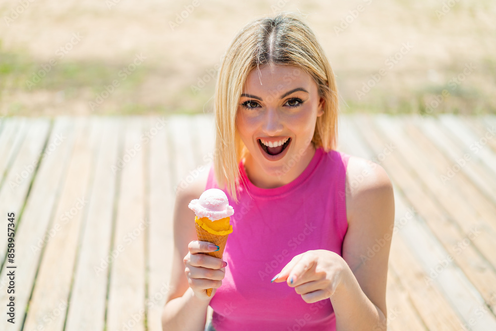 Young pretty blonde woman with a cornet ice cream at outdoors surprised and pointing front Young pretty blonde woman with a cornet ice cream at outdoors surprised and pointing front