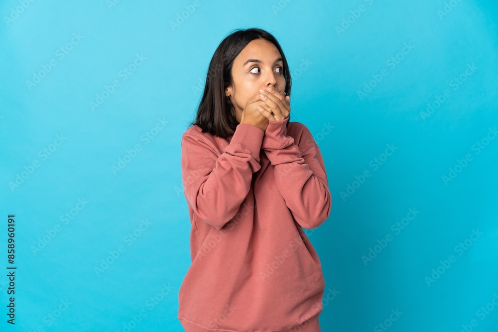 Young latin woman isolated on blue background covering mouth and looking to the side