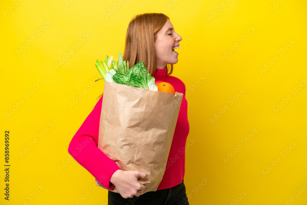 Young redhead woman holding a grocery shopping bag isolated on yellow background laughing in lateral position Young redhead woman holding a grocery shopping bag isolated on yellow background laughing in lateral position