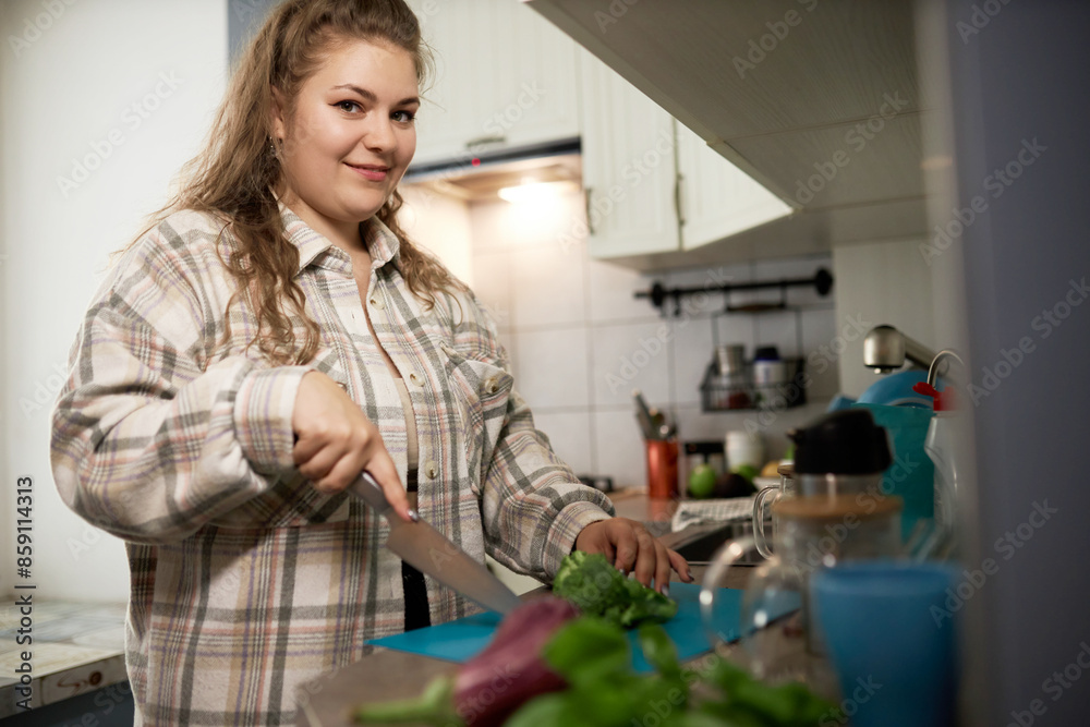 Portrait of plus size young caucasian female chopping vegetables to cook healthy delicious dinner to lose weight being on diet, standing in kitchen with knife and looking at camera with happy face Portrait of plus size young caucasian female chopping vegetables to cook healthy delicious dinner to lose weight being on diet, standing in kitchen with knife and looking at camera with happy face