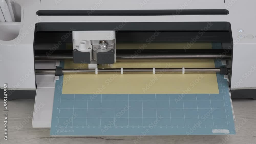 Front view White Smart Cutting machine for home using. Plotter, digital flatbed cut out details on yellow cardboard on blue mat. DIY manual paper craft, making gift for family. Creative Hand crafting Front view White Smart Cutting machine for home using. Plotter, digital flatbed cut out details on yellow cardboard on blue mat. DIY manual paper craft, making gift for family. Creative Hand crafting