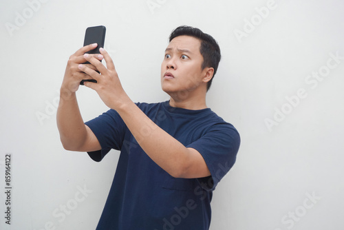 Young Asian man looking smartphone with shocked and surprised expression. Male with blue t-shirt showing excited expression isolated on white background