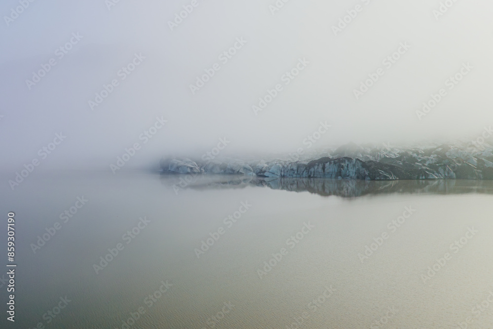 Naklejka premium A serene view of Sólheimajokull glacier in Iceland, shrouded in mist and reflecting in the calm glacial lagoon.