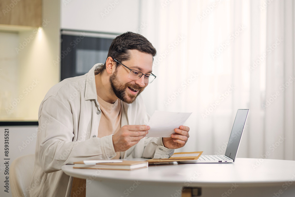 Smiling young man holding paper bill letter while working at table with portable laptop indoors. Skilled caucasian freelancer feeling shock while reading received information at home interior. Smiling young man holding paper bill letter while working at table with portable laptop indoors. Skilled caucasian freelancer feeling shock while reading received information at home interior.