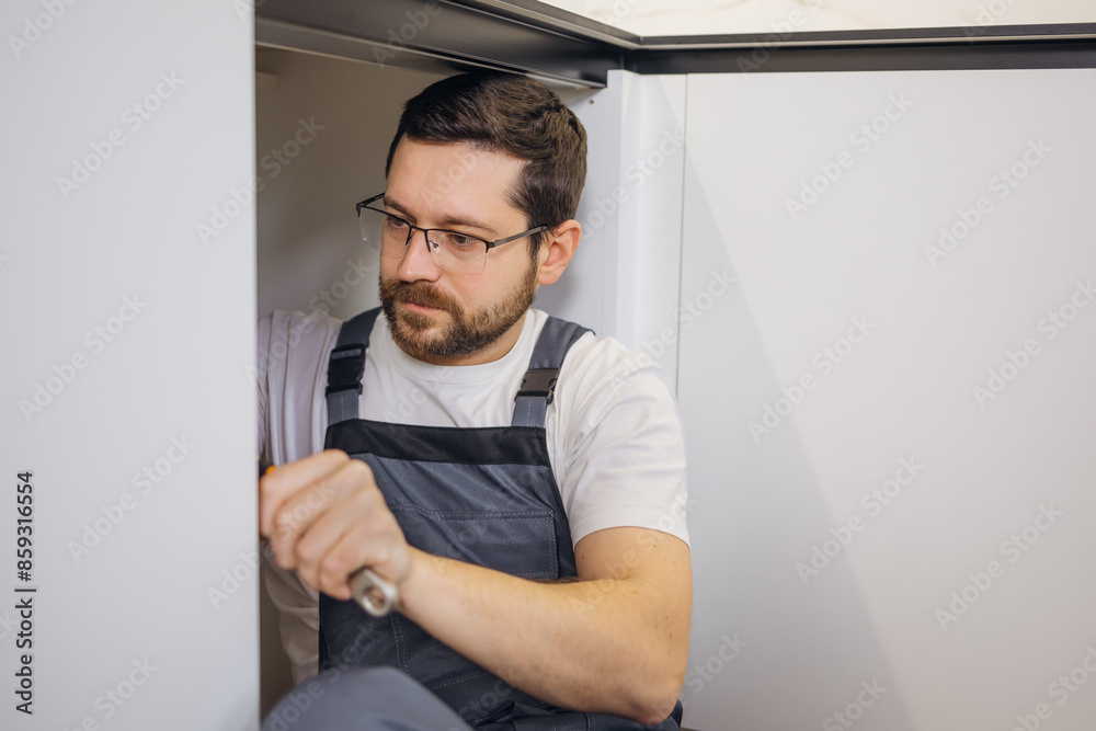 Fototapeta premium Calm caucasian plumber in grey coveralls securely attaching pipe details with screwdriver for long term service. Concerned repairman working with equipment indoors and installing sink parts.