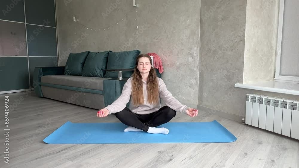 Woman meditates on a yoga mat in a peaceful living room, eyes closed, embracing tranquility. Cross legged pose promotes relaxation and mindfulness