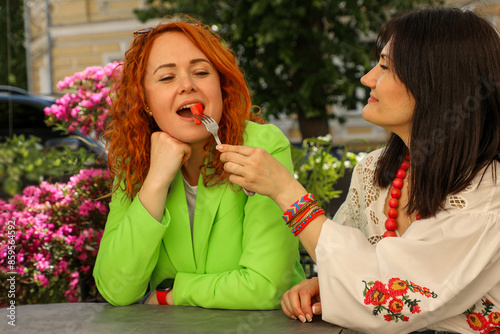 Two women in a cafe are happily eating strawberries, one of them feeds the other with a fork. © Svitlana