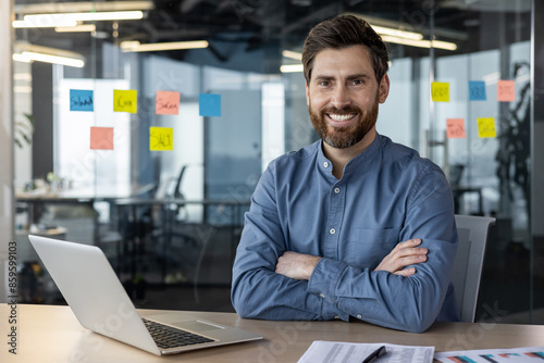 Portrait of a young successful office worker sitting at a desk against a background of a transparent notebook with notes, crossing his arms on his chest and smilingly looking at the camera