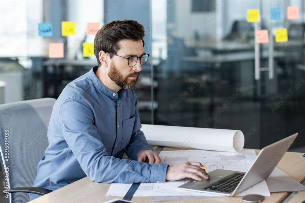 A serious and focused young man, an architect, surveyor and engineer, works in an office with desks and a laptop. Photo from the side A serious and focused young man, an architect, surveyor and engineer, works in an office with desks and a laptop. Photo from the side