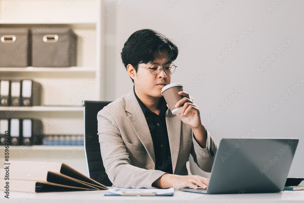 Asian businessmen wearing glasses Sipping coffee while sitting on the computer screen. Concept is to sit and work smart Happy Bright and Relaxed atmosphere. Background is a document rack. Asian businessmen wearing glasses Sipping coffee while sitting on the computer screen. Concept is to sit and work smart Happy Bright and Relaxed atmosphere. Background is a document rack.