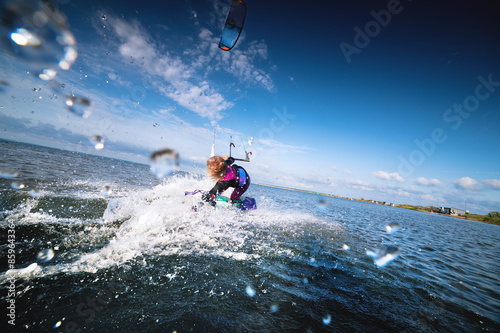 Attractive young Caucasian female kiteboarder doing a trick at sunset with splashes and a smile. Wide angle. Deep dive