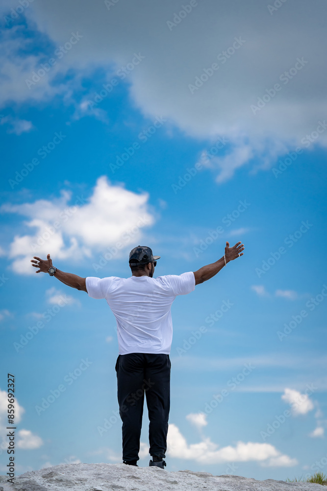 Man Standing On Hilltop With Arms Outstretched. A man in a white t-shirt and black pants stands on a hilltop with his arms outstretched, looking up at the blue sky with white clouds. Man Standing On Hilltop With Arms Outstretched. A man in a white t-shirt and black pants stands on a hilltop with his arms outstretched, looking up at the blue sky with white clouds.
