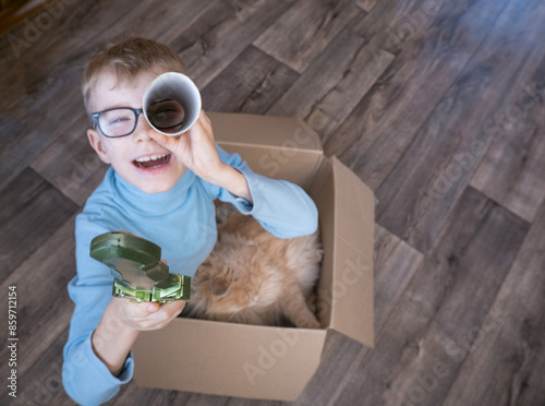 Obraz na płótnie Playful cheerful child, boy looking into a cardboard tube, sitting in a box, playing with a cat w salonie