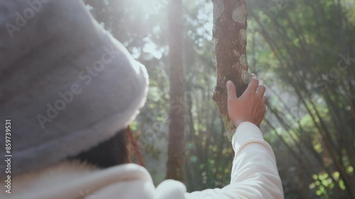 Asian woman wearing warm hat touching a tree trunk in the rainforest with sunlight in the morning time.