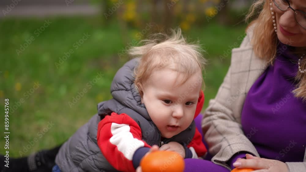 Slow motion. Close-up. A little boy holds out a tangerine, sitting next to his mother on the grass in the park on a spring afternoon. The mother looks at her little son and gently caresses his hair on Slow motion. Close-up. A little boy holds out a tangerine, sitting next to his mother on the grass in the park on a spring afternoon. The mother looks at her little son and gently caresses his hair on