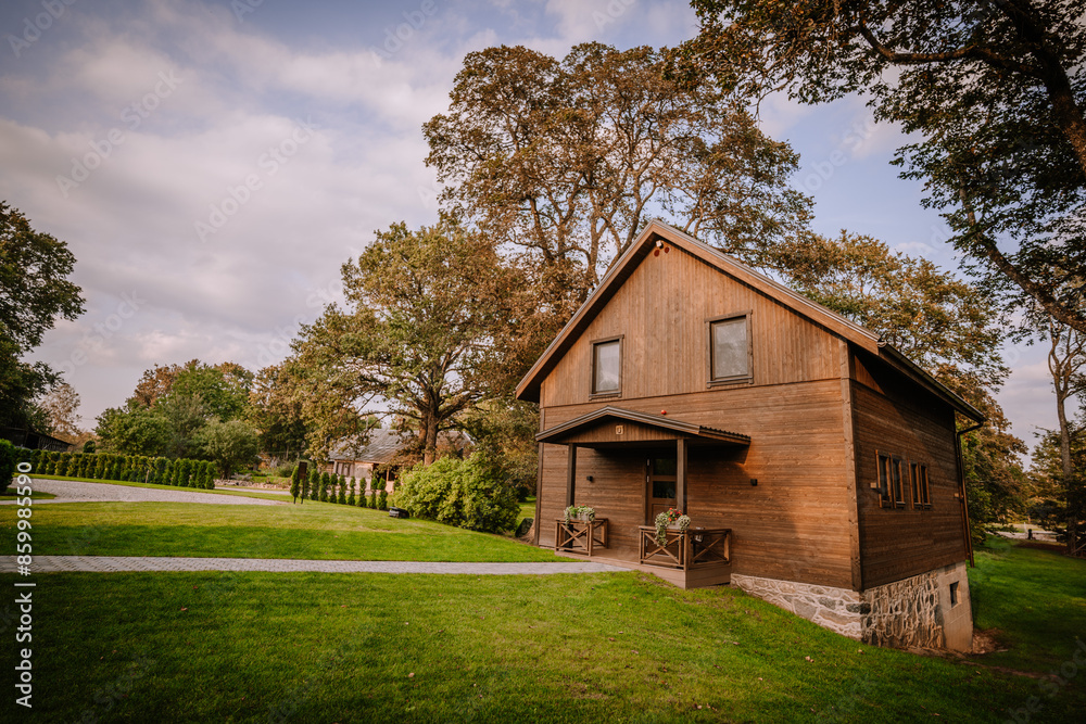 Blome, Latvia - September 11, 2023 - Rustic wooden house surrounded by lush green lawn, trees, and a stone pathway under a partly cloudy sky, creating a serene countryside scene. Copy space. Blome, Latvia - September 11, 2023 - Rustic wooden house surrounded by lush green lawn, trees, and a stone pathway under a partly cloudy sky, creating a serene countryside scene. Copy space.
