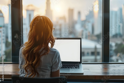 A woman working on her laptop with a blank white screen sitting at an cafe, with a street view of the city in the background, with a bokeh effect, sunlight coming through the window...