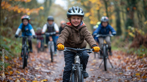 Young boy smiling and leading a group of children on a bike ride through a forest trail covered in autumn leaves.