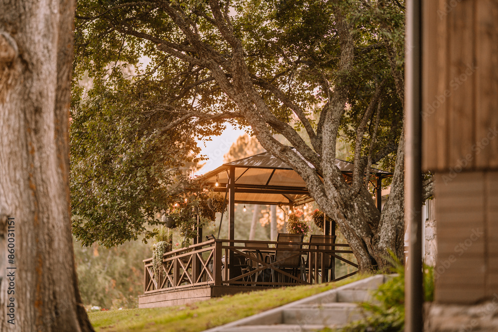 Blome, Latvia - September 11, 2023 - Cozy gazebo nestled among trees at sunset, with hanging plants and outdoor furniture, creating a serene and inviting atmosphere. Copy space. Blome, Latvia - September 11, 2023 - Cozy gazebo nestled among trees at sunset, with hanging plants and outdoor furniture, creating a serene and inviting atmosphere. Copy space.