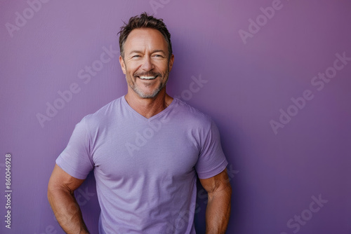 A happy mature man with hands on hips, smiling broadly, dressed in a purple T-shirt, standing against a plain purple backdrop