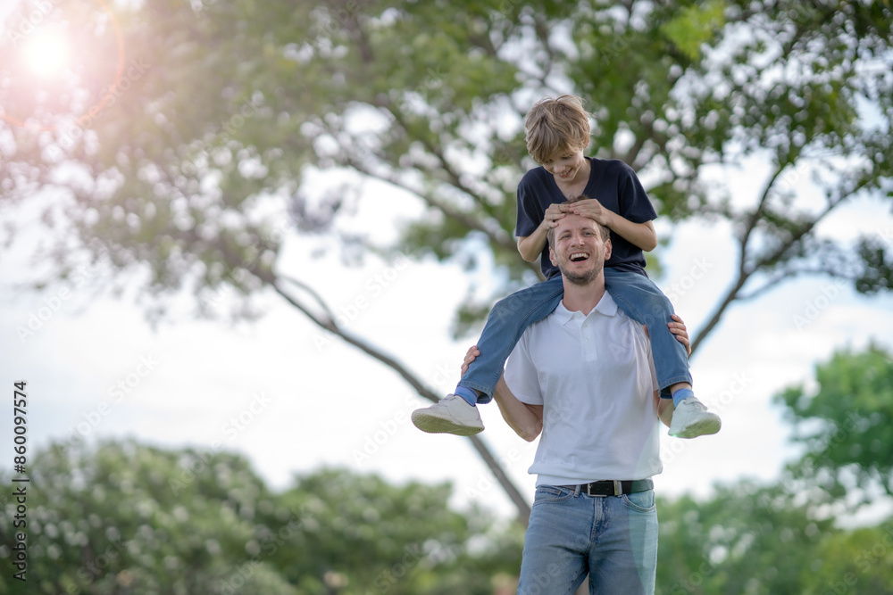 Fototapeta premium White son sitting on his father shoulder with smile and fun, looking his father with love walking in park with blur background and big tree and copy space, relationship between dad and son.