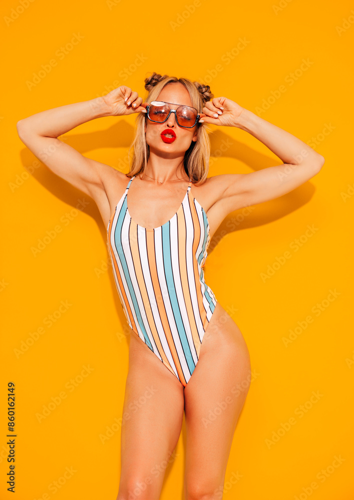 Young smiling blond model in summer swimwear colourful bathing suit. Sexy carefree woman having fun and going crazy. Female posing near yellow wall in studio. Cheerful and happy in sunglasses Young smiling blond model in summer swimwear colourful bathing suit. Sexy carefree woman having fun and going crazy. Female posing near yellow wall in studio. Cheerful and happy in sunglasses