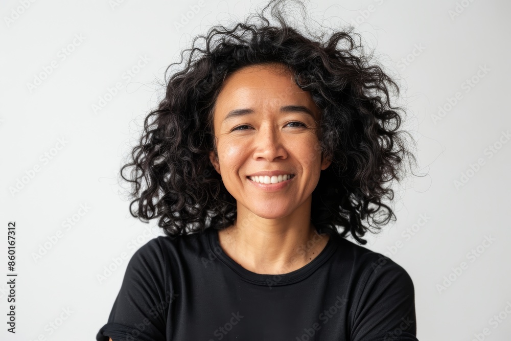 A portrait of a woman with curly, black hair smiling at the camera. She is wearing a black t-shirt and is photographed against a plain white background A portrait of a woman with curly, black hair smiling at the camera. She is wearing a black t-shirt and is photographed against a plain white background
