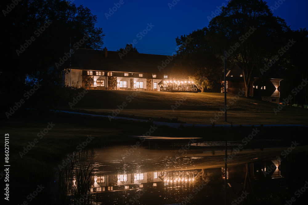 Blome, Latvia - September 11, 2023 - A large, illuminated building at night reflects on a calm pond, creating a serene and picturesque scene. Blome, Latvia - September 11, 2023 - A large, illuminated building at night reflects on a calm pond, creating a serene and picturesque scene.