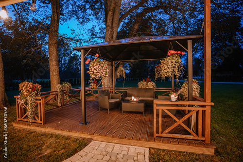 Blome, Latvia - September 11, 2023 - Outdoor patio with wicker furniture, hanging flowers, and a gazebo at dusk, surrounded by trees and ambient lighting, creating a cozy atmosphere. photo