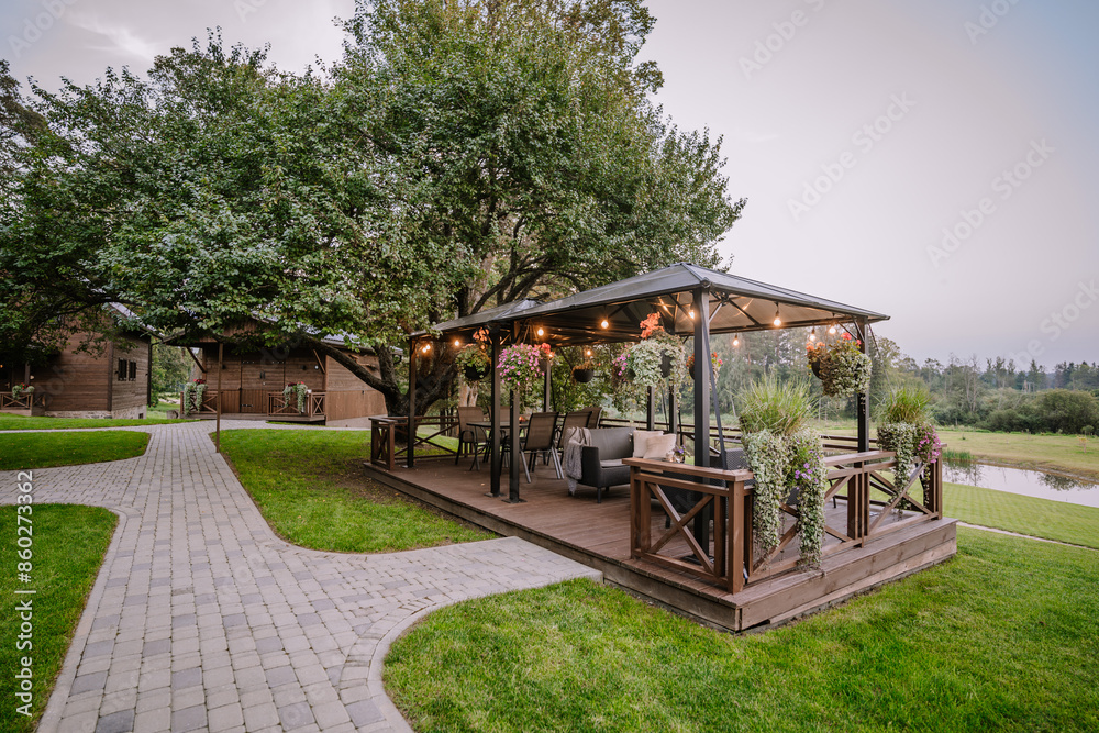 Blome, Latvia - September 11, 2023 - A cozy outdoor patio with string lights, hanging plants, and comfortable seating, set on a lush green lawn with pathways and surrounded by trees... Blome, Latvia - September 11, 2023 - A cozy outdoor patio with string lights, hanging plants, and comfortable seating, set on a lush green lawn with pathways and surrounded by trees...
