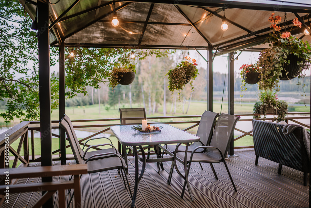 Fototapeta premium Blome, Latvia - September 11, 2023 - A cozy outdoor patio with hanging plants, string lights, and a table set with a candle, offering a serene view of the surrounding greenery.
