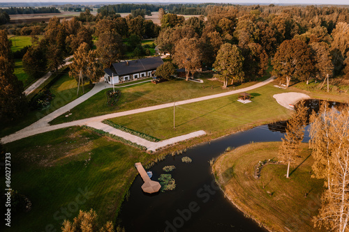 Blome, Latvia - September 11, 2023 - Aerial view of a rural property with a house, lawn, pond with a small dock, gazebo, sandy area, and surrounding trees on a sunny day.