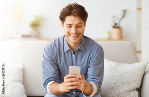 A man smiles while looking at his phone while sitting on a couch.