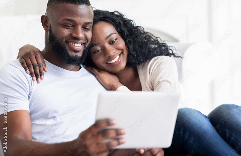 A young couple smiles and relaxes together on a couch while looking at a tablet. A young couple smiles and relaxes together on a couch while looking at a tablet.