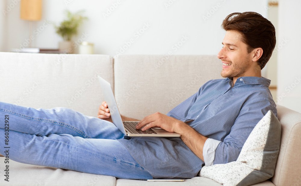 A man is lying on a white couch, using a laptop, while smiling. He is dressed in blue jeans and a blue button-down shirt. A man is lying on a white couch, using a laptop, while smiling. He is dressed in blue jeans and a blue button-down shirt.