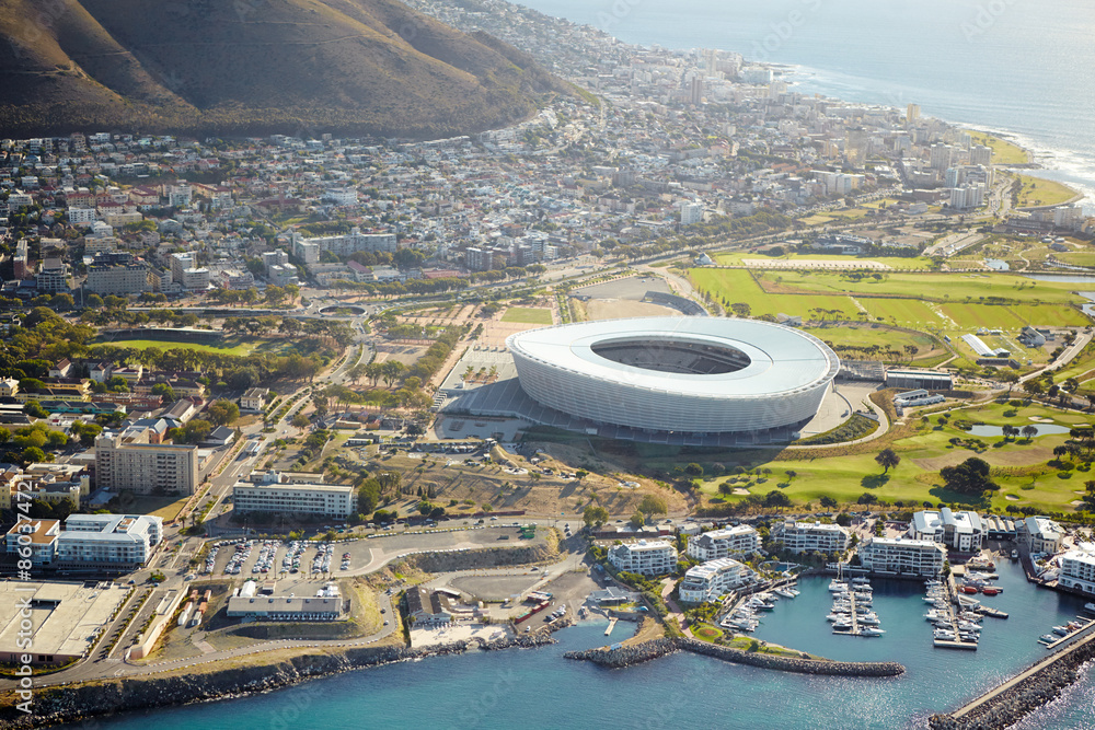 Fototapeta premium Harbor, drone and stadium in city for travel with ships, cruise or buildings by ocean and mountain. Boats, coast and aerial view of infrastructure by seashore for holiday destination in Cape Town.