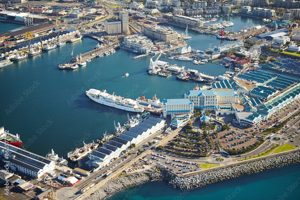 Obraz premium Harbor, drone and buildings by dock in ocean for travel with cargo ships or cruise in city. Infrastructure, coast and aerial view of boats at shore by sea water for holiday destination in Cape Town.