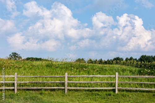 A split-rail fence with grass, trees and cumulus clouds in the background. 