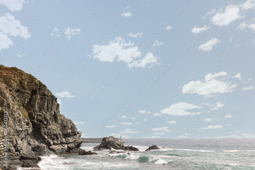 A rocky shoreline with a bridge in the distance. The sky is clear and blue with a few clouds A rocky shoreline with a bridge in the distance. The sky is clear and blue with a few clouds