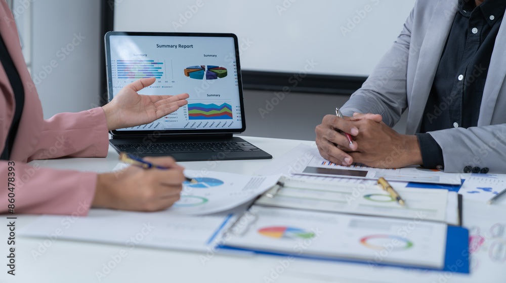 A photograph of a male and female employee's hands as they meet in an office, pens and laptops at the ready as they consult on business matters.