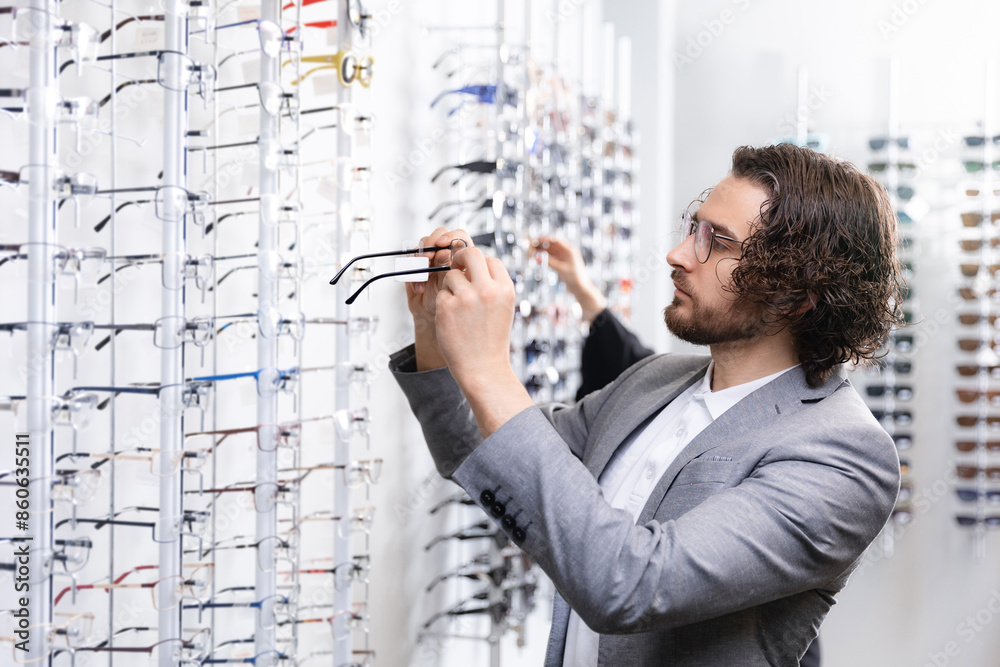 Happy young man chooses spectacle frames in optician shop. Stylish fashion male in glasses at optic store, light background Happy young man chooses spectacle frames in optician shop. Stylish fashion male in glasses at optic store, light background