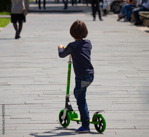 A young boy in jeans and a dark blue shirt starts riding a green scooter in park