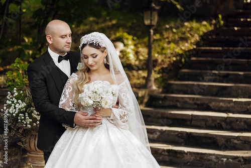 A bride and groom are posing for a picture in front of a staircase. The bride is wearing a white dress and a veil, while the groom is wearing a black suit. They are holding a bouquet of flowers