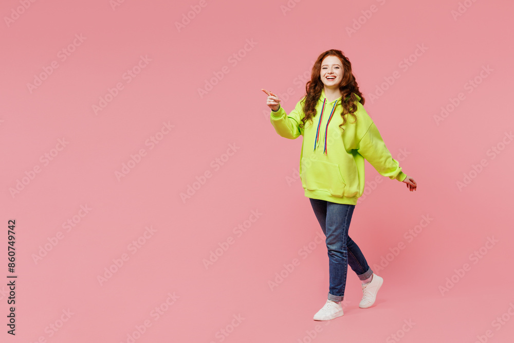 Full body young smiling happy ginger woman wear green hoody casual clothes walk go point index finger aside on area isolated on plain pastel light pink background studio portrait. Lifestyle concept. Full body young smiling happy ginger woman wear green hoody casual clothes walk go point index finger aside on area isolated on plain pastel light pink background studio portrait. Lifestyle concept.