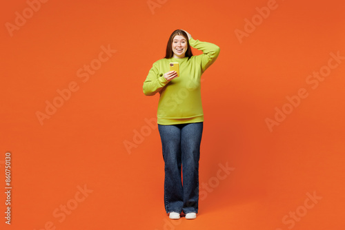 Full body shocked young ginger chubby plus size woman wear green sweatshirt casual clothes hold in hand use mobile cell phone isolated on red orange background studio portrait. Body positive concept. photo